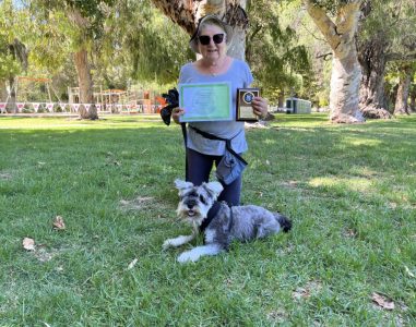 Woman with her dog holding a certificate and award outdoors in a park setting.