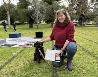 Woman with her dog holding a certificate in a park during an outdoor event.
