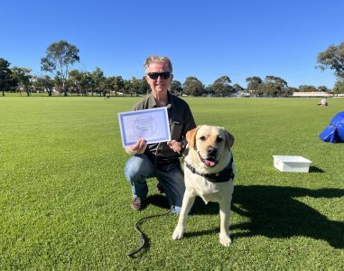 A dog owner holding a certificate with a Labrador Retriever in a park setting.